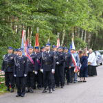 Grupa strażaków w mundurach stoi w szeregu z flagami. W tle zielone drzewa i zaparkowany samochód. Uroczysta atmosfera.