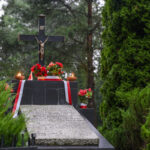 Grób z krzyżem i Chrystusem pośród zielonych drzew. Czerwono-białe kwiaty i wstęga dekorują miejsce. Pochmurne niebo w tle.