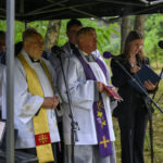 Na zdjęciu kilku księży w białych i ozdobnych szatach, obok kobieta z książką. Tło zielone, z drzewami. Mają mikrofony pod namiotem.
