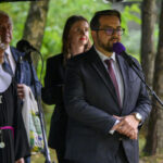 Zdjęcie przedstawia mężczyznę w garniturze przemawiającego do mikrofonu. Obok stoi zakonnica, w tle drzewa. Kolory dominujące to zieleń i szarość.