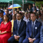 Grupa ludzi siedzi pod zielonym namiotem; na pierwszym planie osoby elegancko ubrane, w tle widoczne kolorowe parasole.