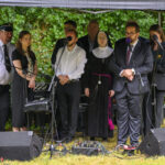 Grupa osób stoi pod namiotem na tle zieleni, przemawiają do mikrofonów. Są ubrani w formalne stroje. Na trawie leżą kable i sprzęt nagłaśniający.