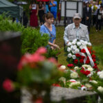 Trzy osoby składają wieniec z czerwonych i białych róż na cmentarzu. Tło jest zielone, z grupą ludzi w oddali. Scena pełna kwiatów.