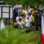 Grupa ludzi z kwiatami stoi na mokrej trawie pod parasolami. Ubrani w białe koszulki, w tle zielone drzewa. Tablica na pierwszym planie.