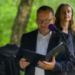 Mężczyzna w okularach czyta z kartki przy mikrofonie na tle zieleni. Obok stoi kobieta. Oboje ubrani są elegancko.