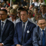 Grupa ludzi w garniturach siedzi w rzędzie na wydarzeniu. W tle widać innych uczestników. Atmosfera jest formalna.