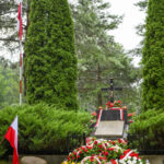 Grób z krzyżem na tle zieleni, otoczony wieńcami biało-czerwonych kwiatów. Flaga Polski po lewej. W tle drzewa.