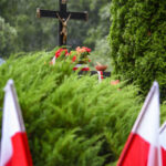 Czarne krzyż z Jezusem na tle zielonych drzew, otoczony czerwonymi kwiatami i polskimi flagami.