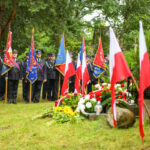 Ceremonia w lesie z uczestnikami w mundurach i flagami w kolorach Polski. W tle zielone drzewa. Na przodzie kwiaty i znicze.