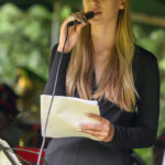 Osoba trzyma mikrofon i notatki, śpiewając. Ubrana na czarno, w tle rozmyta zieleń. Występ na świeżym powietrzu.