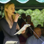 Na zdjęciu kobieta w czarnej sukience trzyma mikrofon i kartki. W tle widoczny jest tłum ludzi, pod zielonym namiotem. Tło jest nieostre, z dominacją zieleni.