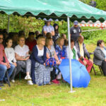 Grupa ludzi siedzi pod zielonym namiotem, na trawie, z otwartym niebieskim parasolem. W tle zielone liście i park. Atmosfera jest spokojna.