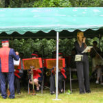 Zespół muzyczny w czerwonych mundurach gra pod zielonym namiotem. Dyrygent stoi tyłem, obok kobieta mówi do mikrofonu. Publiczność siedzi w tle.