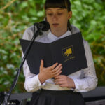 Na zdjęciu młoda osoba czyta z czarnej teczki, nosi białą koszulę. Tło stanowi zielone liście, a osoba stoi przy mikrofonie. Po prawej widać fragment instrumentu klawiszowego.