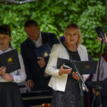 Grupa osób na scenie czyta z teczek. W tle zielone liście. Kolorystyka: biel, czerń, granat.