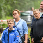 Grupa osób stoi na zewnątrz na tle zieleni. Przeważają niebieskie i czarne ubrania. Tło jest rozmyte, a ogrodzenie w tle ma biało-czerwone taśmy.