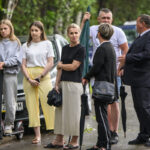 Grupa ludzi stoi na ulicy z zielenią w tle. Jedna osoba trzyma hulajnogę, inna ma czarną parasolkę. Kobieta w czarnym swetrze nosi torbę na ramieniu.