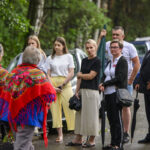Grupa ludzi stoi na ulicy, w tle drzewa. Dwie osoby w czerwonych chustach z kwiatami. Kilka osób trzyma parasole.