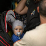 Na zdjęciu widoczny jest niemowlak w czapce, z smoczkiem, siedzący w wózku o ciemnym kolorze z czerwonymi akcentami. Obok dziecka znajduje się dorosły. Tło jest rozmyte, ciemne.