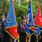 Strażacy w czarnych mundurach stoją z kolorowymi sztandarami: niebieskim i czerwonym. W tle zielone drzewa.