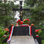 Czarny krzyż z figurą Jezusa na tle zielonych drzew. Przed krzyżem czerwone kwiaty i biało-czerwone wstążki. Przy krzyżu płoną znicze.