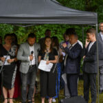 Grupa ludzi pod czarnym namiotem, trzymają mikrofony i kartki. W tle zielone drzewa. Uczestnicy w eleganckich strojach.