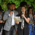 Grupa osób śpiewa na scenie z mikrofonami. Tło z zielonymi liśćmi. Ubrani są elegancko, w ubrania w różnych kolorach.