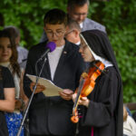 Grupa ludzi stoi na scenie z mikrofonami, jedna osoba trzyma skrzypce. W tle zielone liście. Kolorystyka stonowana.
