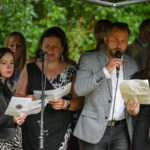 Grupa osób śpiewa z nutami w rękach. Są na zewnątrz, otoczeni zielonymi drzewami. Mają czarno-białe ubrania.