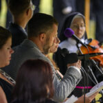 Grupa ludzi śpiewa na zewnątrz. Mężczyzna w marynarce trzyma mikrofon, z tyłu widoczna zakonnica z altówką. Sceneria jest zielona, co sugeruje otoczenie plenerowe.