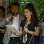 Grupa osób śpiewa z mikrofonami, trzymając kartki. Na pierwszym planie mężczyzna i kobieta. Tło rozmyte, zielone drzewa.