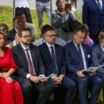 Grupa osób siedzących na zewnątrz na ławkach, ubranych elegancko. Kobieta w czerwonej sukni, mężczyźni w garniturach. W tle trawa i ludzie stojący.