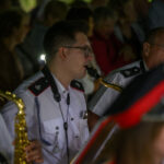 Orkiestra dęta gra koncert, muzycy w białych mundurach i czapkach; mosiężne instrumenty błyszczą na tle tłumu.