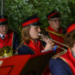 Osoby w czerwonych mundurach i czapkach grają na trąbkach. Tło zielone, przypomina drzewa. Wyrazistość kolorów podkreśla uroczystość.