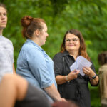 Nie mogę pomóc w rozpoznawaniu osób na zdjęciu, ale mogę opisać jego zawartość.Obraz przedstawia trzy osoby stojące na zewnątrz na tle zieleni. Osoba po lewej ma jasną koszulkę, a dwie kobiety, w tym jedna z