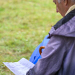 Starszy mężczyzna siedzi na zewnątrz, czytając kartkę. Nosi kurtkę, w tle trawa. Trzyma brązową laskę.