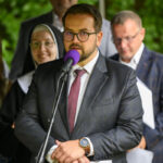 Mężczyzna w garniturze przemawia do mikrofonu, obok grupa ludzi. Tło jest zielone. Kobieta w habicie uśmiecha się.