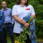 Kobieta w białej koszulce trzyma słoneczniki i kurtkę jeansową. W tle zielone drzewa i dwie osoby. Jest na zewnątrz na chodniku.