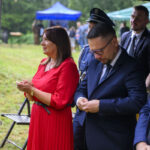 Grupa ludzi na zewnątrz, kobieta w czerwonej sukience, mężczyźni w garniturach. W tle zielona trawa i polskie flagi.