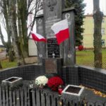 Pomnik z szarym krzyżem i polskimi flagami. Obok biało-czerwone chryzantemy. Tło to drzewa i ogrodzenie.