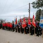 Grupa strażaków stoi w szeregu z czerwonymi i niebieskimi sztandarami. Ubrani są w czarne mundury i czerwone kurtki. Tło stanowi budynek z czerwonym dachem i drzewa.