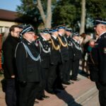 Grupa osób w mundurach strażackich stoi na chodniku. Obok nich kilka osób w cywilnych ubraniach. W tle widać drzewa i budynek.