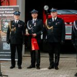 Czterech strażaków w ciemnych mundurach stoi przed czerwonym wozem strażackim. Dwóch z nich trzyma nagrody. W tle jest transparent OSP.