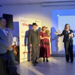 Na zdjęciu widać cztery osoby na jasnym tle. Trzy z nich stoją na scenie, dwie trzymając mikrofon. Tło zdobi napis Mazowsze.