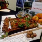 Na stole ciasto z ziołami i papryką, obok jabłka i orzechy. W tle ciemny obrus, ozdoby z liści i plakat Mazowsza.