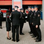 Grupa strażaków w czarnych mundurach stoi przed remizą z czerwonymi drzwiami. W tle widoczny jest czerwony wóz strażacki. Strażacy uczestniczą w ceremonii na placu z kostką brukową.