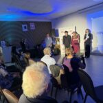 Grupa ludzi w sali konferencyjnej na tle niebieskiej ściany. Mężczyzna i kobieta w eleganckich strojach na scenie. Publiczność siedzi przodem do sceny, niektóre osoby robią zdjęcia.