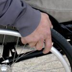 Osoba w szarej kurtce trzyma wózek inwalidzki. Tło jest rozmyte, dominuje szarość i czerń. Ręka skupiona na metalowym kole.