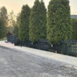 Wzdłuż asfaltowej drogi znajduje się czarny metalowy płot. W tle są wysokie zielone drzewa. Niebo ma ciepły odcień zachodu słońca.