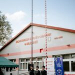 Czterech strażaków stoi przed budynkiem straży pożarnej z biało-czerwonymi flagami. Budynek jest szary z czerwonymi detalami. Tło stanowi niebieskie niebo z chmurami.